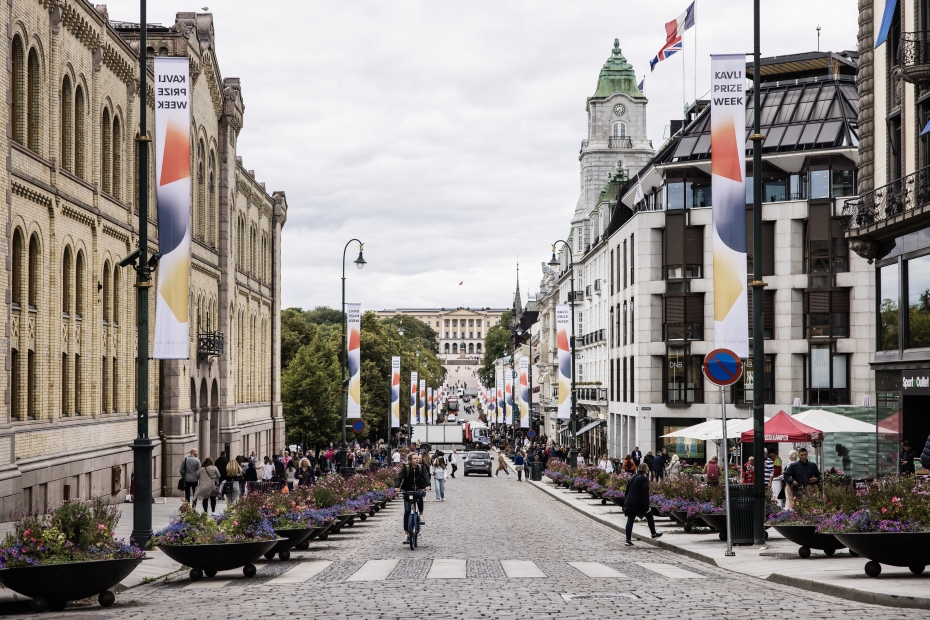The main street of Oslo, Karl Johan avenue, is decorated with The Kavli Prize banners. They are white with the blue circle, yellow hectagon, and the red triangle. The street is busy. The palace is seen at the end. 