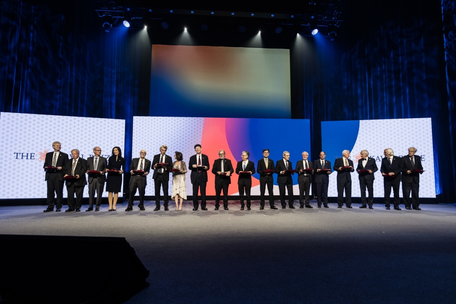 18 Kavli Prize laureates on stage of Oslo Concert House. They hold their Prize scroll, containing the prize and the official diploma. They are all wearing suits or dresses. They look happy. 