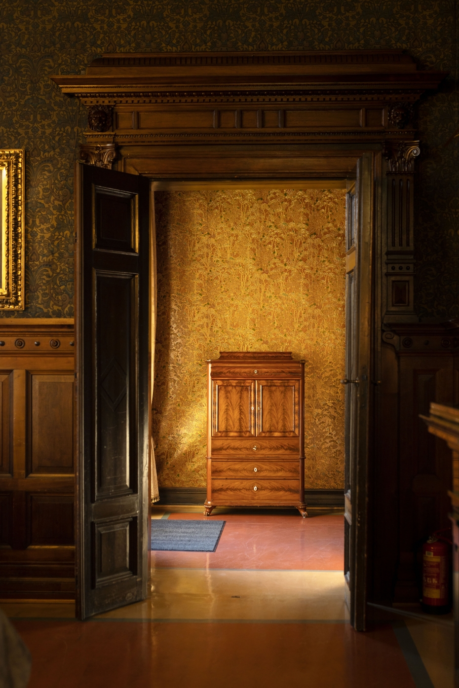 Stor dør med utforming i mørkt tre åpnes til et gullbelagt kabinettrom med solskinn. 